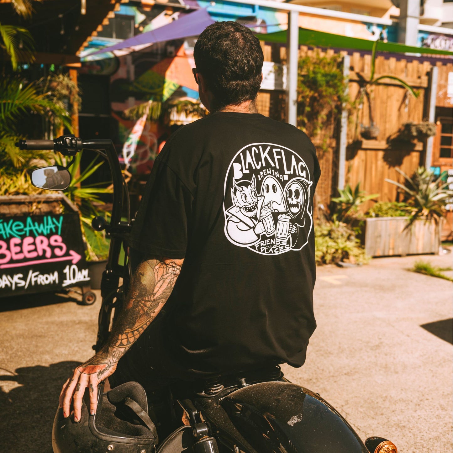 Man on motorcycle wearing a black Blackflag t-shirt with hand drawn Friends In Low Places illustration on back.